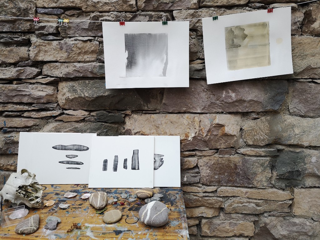 Works on paper clipped against a stone wall with found objects below.